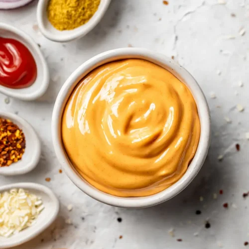 Top view of a bowl of creamy orange burger sauce surrounded by sliced red onions, mustard powder, ketchup, red pepper flakes, shredded cheese, and part of a burger bun on a white textured background.