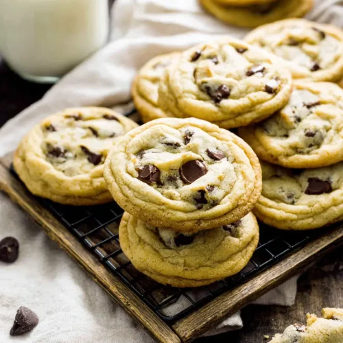 Stack of soft, homemade chocolate chip cookies on a cooling rack with scattered chocolate chips and a glass of milk in the background.