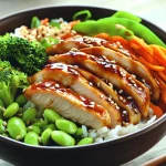 Teriyaki chicken rice bowl topped with sliced glazed chicken, sesame seeds, white rice, edamame, shredded carrots, snap peas, and steamed broccoli.