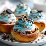 Cookie Monster cinnamon roll topped with swirls of blue frosting, crushed Oreo pieces, and cookie crumbles on a white plate.