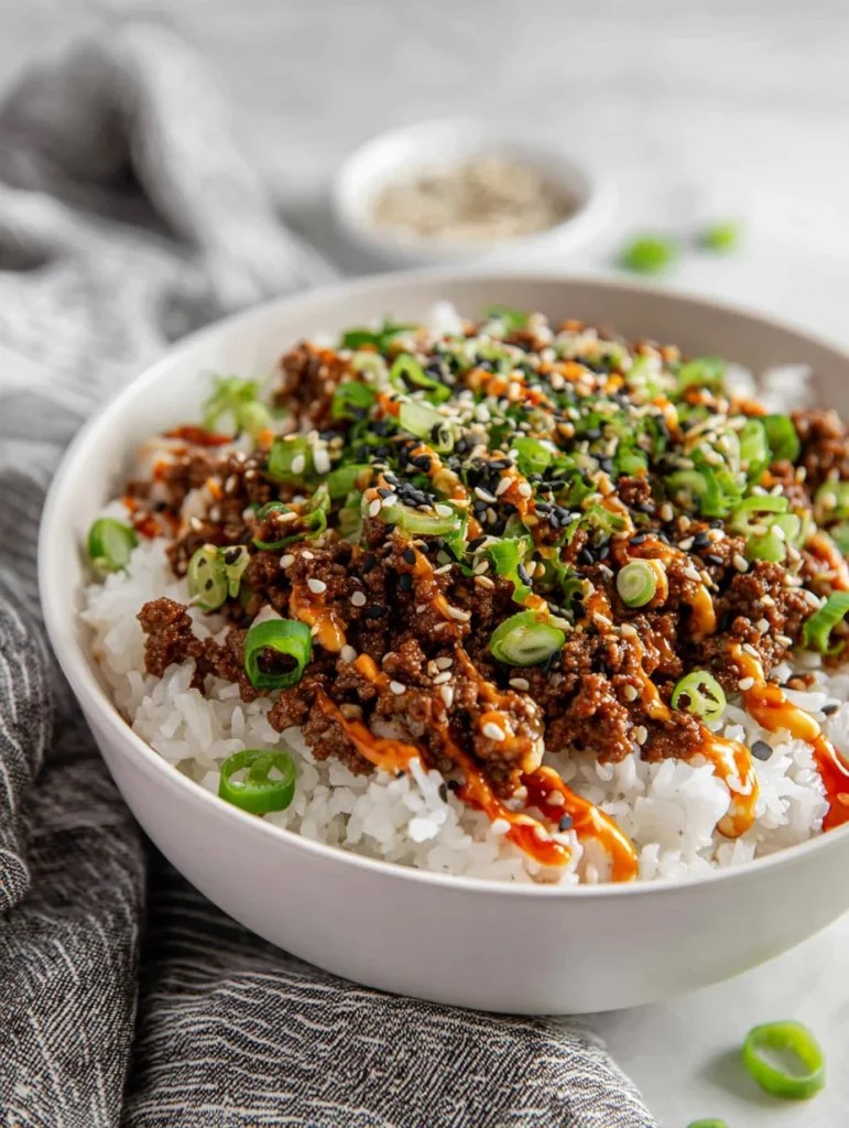 Korean ground beef bowl with white rice, drizzled with spicy sauce and topped with sesame seeds and chopped green onions.