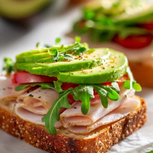 Open-faced turkey avocado sandwich topped with sliced avocado, fresh arugula, tomato, and deli-sliced turkey on whole-grain bread.